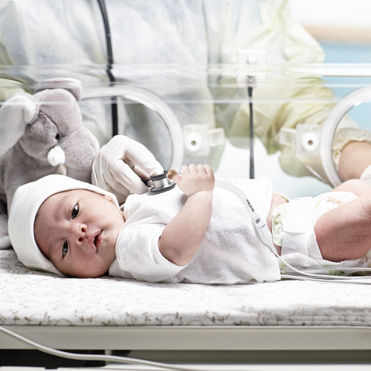 Bebé en la incubadora de la unidad de cuidados intensivos neonatales examinado por un médico con un estetoscopio y monitorizado con un sensor de oximetría de pulso Masimo RD SET.